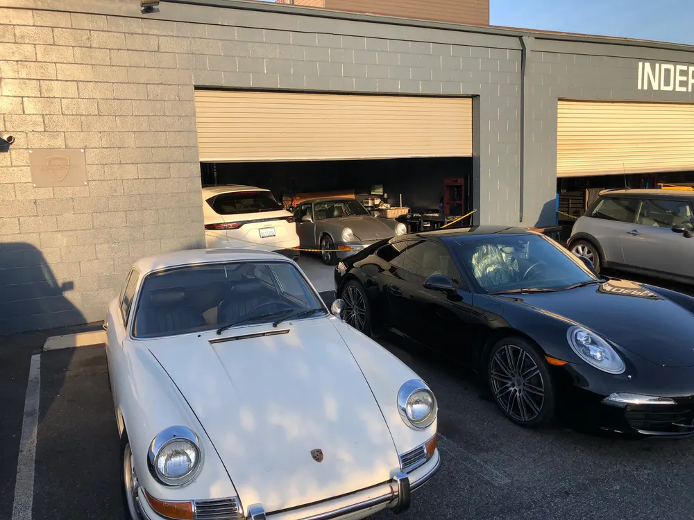 White classic Porsche and a black modern 911 parked outside the shop's bay door